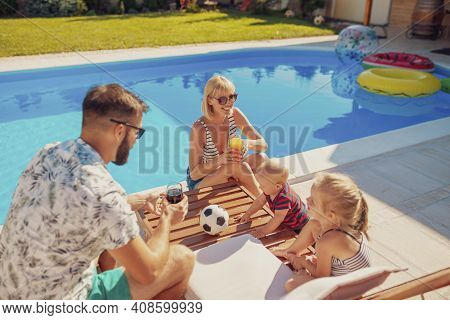 Beauitful Young Family Spending Hot Sunny Summer Day By The Swimming Pool, Parents Relaxing And Drin