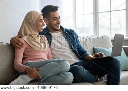Relaxed Middle-eastern Couple Sitting On Couch At Home, Using Laptop, Smiling Bearded Arab Man And W