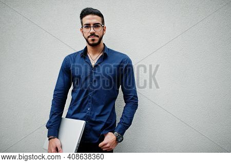 Middle Eastern Entrepreneur Wear Blue Shirt, Eyeglasses, Hold Laptop Against White Wall.
