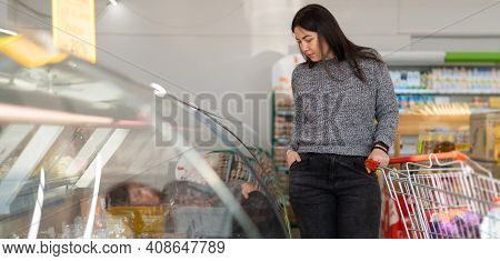 A Cute Brunette Caucasian Girl Stands In The Supermarket In The Department With Products In Refriger