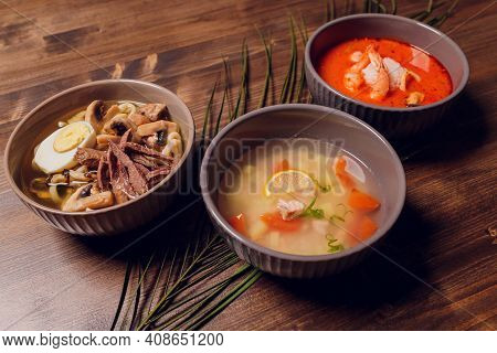 Set Of Serving Asian Soups On Gray Background Top View. Tom Yum Soup, Eel Fish Soup And Ramen Noodle