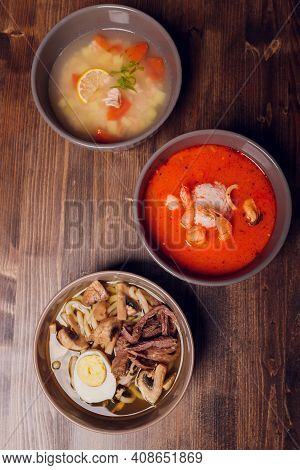 Set Of Serving Asian Soups On Gray Background Top View. Tom Yum Soup, Eel Fish Soup And Ramen Noodle