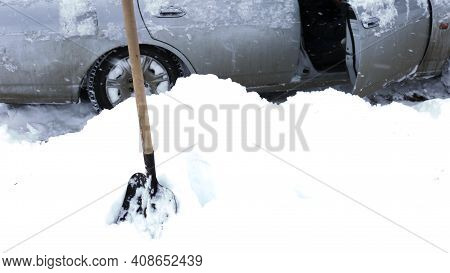 A Snow Shovel Stuck In A Snowdrift On The Side Of The Road Against The Background Of A Car Caught In