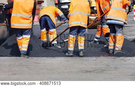 A Team Of Road Workers In Orange Reflective Uniforms Repair A Section Of Road With Fresh Asphalt. Cl