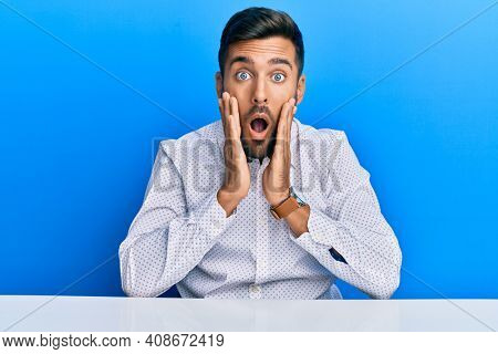 Handsome hispanic man wearing business clothes sitting on the table afraid and shocked, surprise and amazed expression with hands on face 