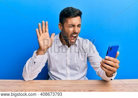 Handsome hispanic man doing video call waving to smartphone angry and mad screaming frustrated and furious, shouting with anger. rage and aggressive concept. 