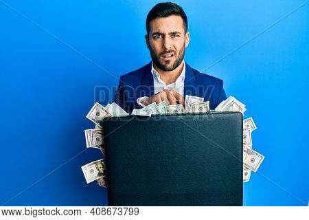 Young hispanic businessman holding briefcase full of dollars clueless and confused expression. doubt concept. 
