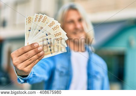 Young blond scandinavian man smiling happy holding danmark 100 kroner banknotes at the city.