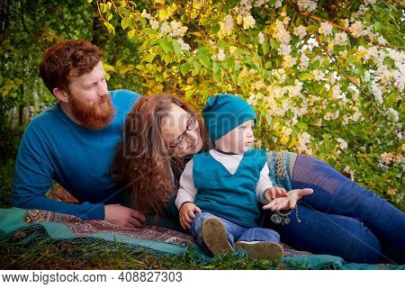 Young Family With Kid Having Picnic Outdoors Near Apple Tree With White Flowers. Parents With One Ch