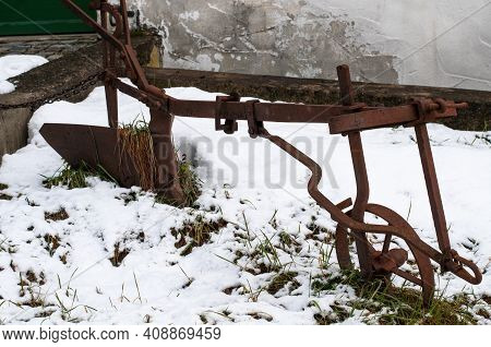 An Old Plough Covered With Rust As Decoration In A Sowy Garden
