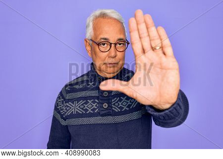 Middle age senior grey-haired man wearing glasses and winter sweater over purple background doing stop sing with palm of the hand. Warning expression with negative and serious gesture on the face.