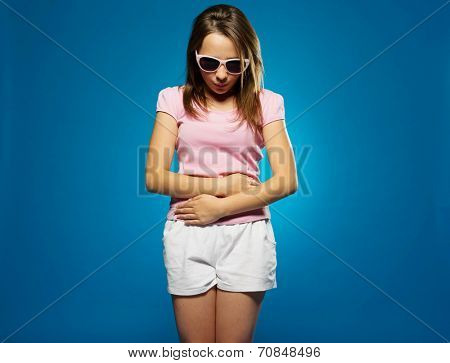 Young girl with a stomach ache clutching her belly with her hands looking down dejectedly at the floor, on blue
