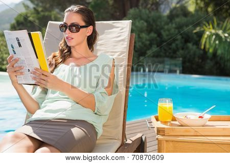 Beautiful young woman reading book by swimming pool with breakfast on table