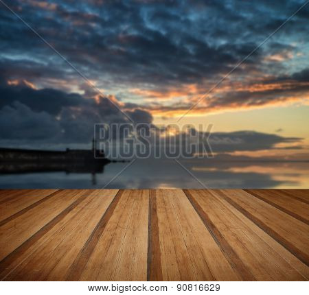 Beautiful Vibrant Sunrise Sky Over Calm Water Ocean With Lighthouse And Harbor Wall With Wooden Plan