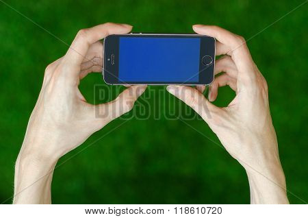 Human Hand Holding A Modern Mobile Phone With A Blue Screen On A Background Of Green Grass First-per