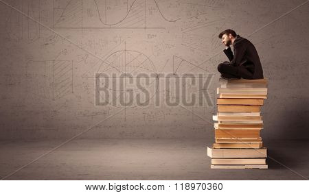 A serious businessman in suit sitting on a pile of giant books in front of a greyish brown wall with drawn lines, angles, numbers, circles and curves.