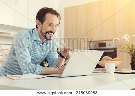 Funny picture. Happy cheerful positive man looking at the laptop screen and laughing while pointing at it