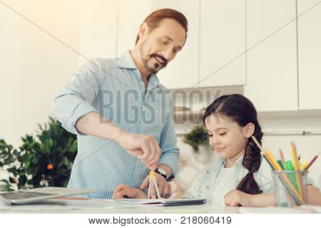 Geometry lesson. Joyful nice intelligent man standing at the table and using a pair of compasses while having a geometry lesson with his daughter