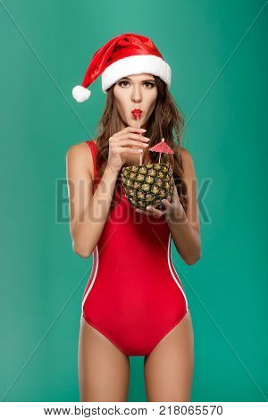 beautiful woman in red bikini and christmas hat drinks summer cocktail in pineapple. christmas holiday