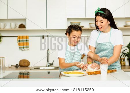 daughter helps her mother knead the dough , standing at white kitchen at home and laughing . Cooking together with kid
