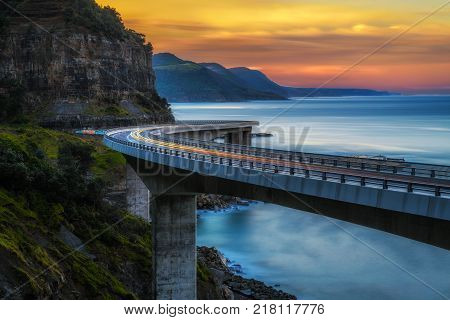 Sunset over the Sea cliff bridge along Australian Pacific ocean coast with lights of passing cars near Sydney, Australia. Long exposure.