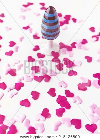 Ceramic butt plug with a pattern in blue and black stripes is on a white background with red and pink hearts made of cloth. Romantic background for Valentine's Day or for sex shop advertising