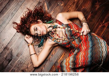 Beautiful bright woman with curly foxy hair wears a dress and beads in boho style, lies on the wooden floor. Ethnic style in clothes.