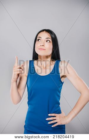 young thoughtfull teen with long brunette hair in blue dress, holding finger up in photostudio on gray background