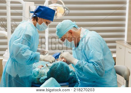 Male Old Professional Dentist Surgeon In Uniform And Female Assistant Helps To Performing Operation