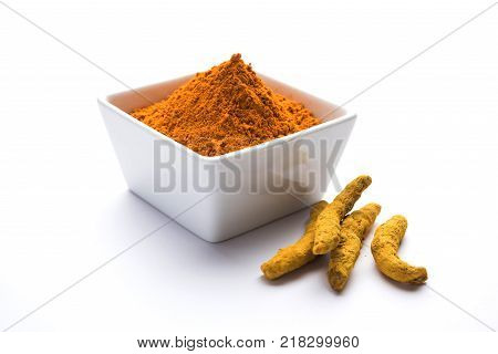 Turmeric powder in ceramic bowl with raw dried turmeric over plain background
