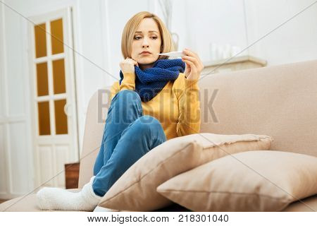 Having fever. Pretty upset young blond woman feeling in the blues and holding a thermometer and having a blue warm scarf and a yellow sweater on while sitting on the couch