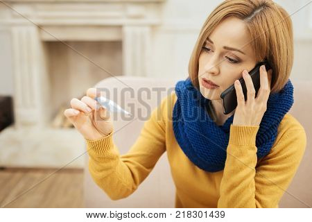 Fever. Upset young blond woman feeling bad and holding a thermometer and having a blue warm scarf on while sitting on the couch and talking on the phone
