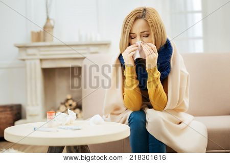 Under the weather. Ill young blond woman feeling bad and blowing her nose while having a blanket on her shoulders and sitting on the couch with her eyes closed and table with pills in front of her