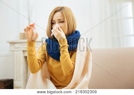 Being sick. Unhappy unwell young blond woman having fever and blowing her nose and holding medication while having a blanket on her shoulders and sitting on the couch