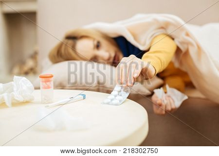 Drug regimen. Sick young blond woman feeling under the weather and lying on the couch and talking pills from the table situating in front of the couch