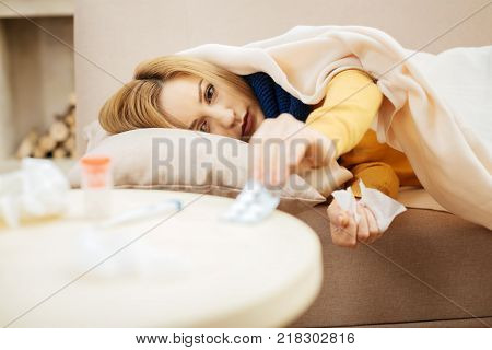 Medication administration. Seedy young blond woman feeling under the weather and lying on the couch and talking pills from the table situating in front of the couch