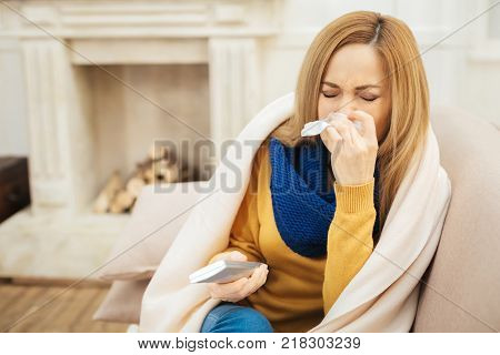 Sneezing. Sick unhappy young dark-eyed woman having runny nose and sneezing and sitting on the couch and holding remote control and a fireplace in the background