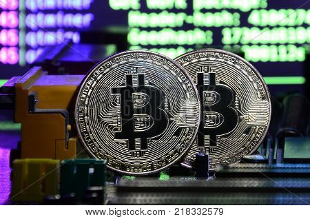 Two bitcoins lies on a videocard surface with background of screen display of cryptocurrency mining by using the GPUs