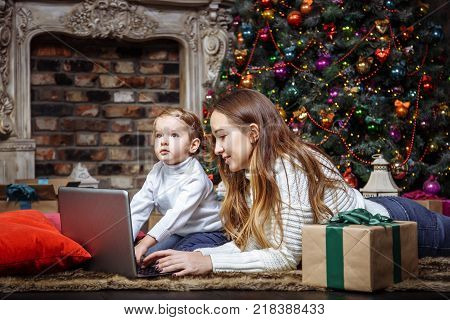 Happy young family mother and daughter using a laptop while sitting near the Christmas tree.