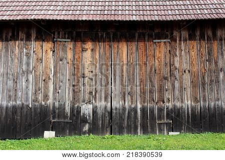 Large unpainted wooden barn doors with strong metal hinges, red roof tiles and uncut grass in front on warm sunny day