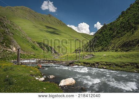 Landscape of a mountain valley with view at mountain river and mountain range. Blue sky with clouds over mountains. Bridge over a mountain river.