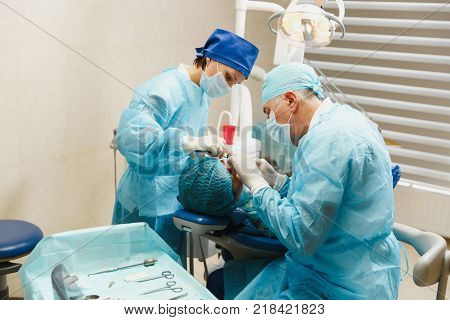 Male Old Professional Dentist Surgeon In Uniform And Female Assistant Helps To Performing Operation