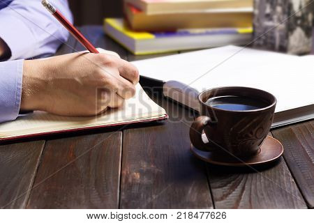 Male hand holding notepad or book with white clean pages black coffee in brown cup red pencil on dark wood working table working or study concept.