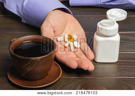 Male hand holds vitamin complex and dietary supplement capsules of omega 3 glucosamine calcium pills cup of black coffee on dark wooden table.
