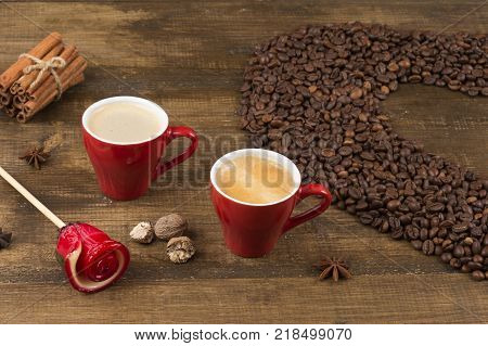 Two cups of coffee with a foam top view. Coffee beans badon cinnamon and caramel on a wooden surface. Love of a coffee drink. Morning invigorating coffee for two. Heart made of coffee beans.