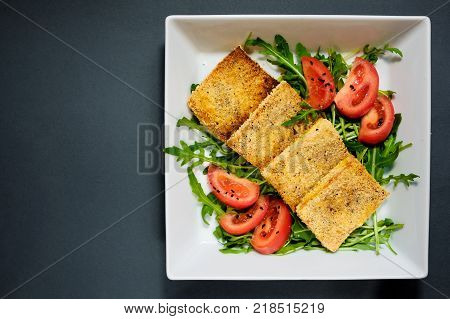 Salad of fried tofu with tomatoes and rocula. Homemade vegetarian salad in a white plate on a black background. A healthy Asian diet vegan vegetarian salad.