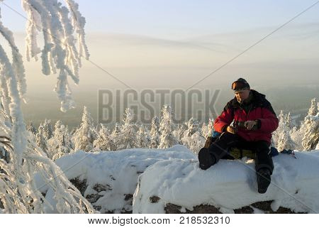 the hiker sitting on a backpack at the top of the rock in winter rests pouring himself a tea from a thermos
