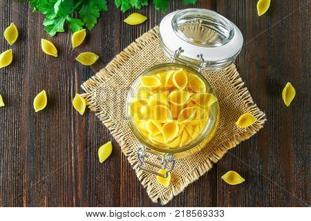 Culinary background with conchiglie pasta on wooden table. Pasta in the form of cockleshells in a glass jar with parsley. Top view.
