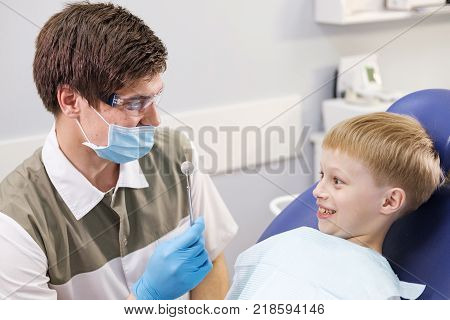 Male dentist examines the teeth of the patient cheerful child with blond hair. Boy smiling in dentist's chair with mouth wide open.