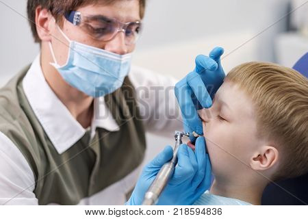 Male dentist examines the teeth of the patient cheerful child with blond hair. Boy smiling in dentist's chair with mouth wide open.